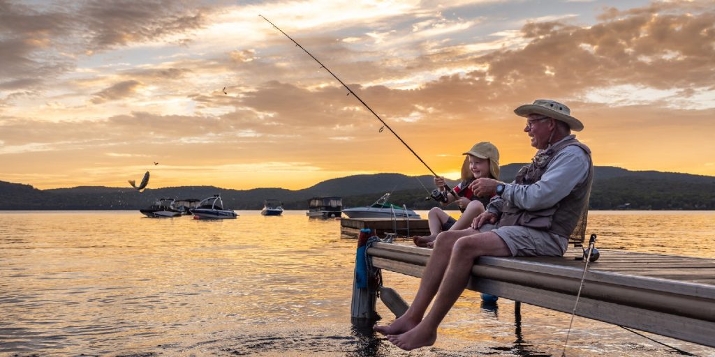 A grandfather is teaching his grandson to fish during sunset in summer.