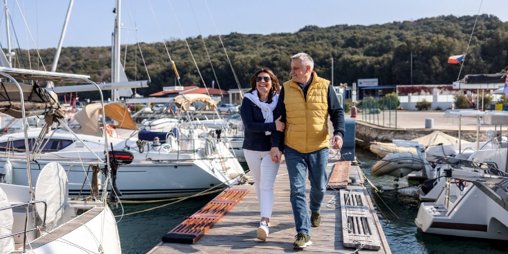 Mature Caucasian couple walking on pier at the harbor, holding their hands and having a great time together.