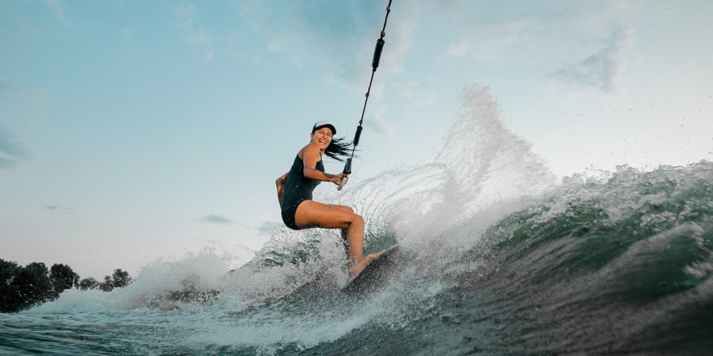 Young beautiful brunette woman riding on the wakesurf holding a rope of a motorboat on the background of trees