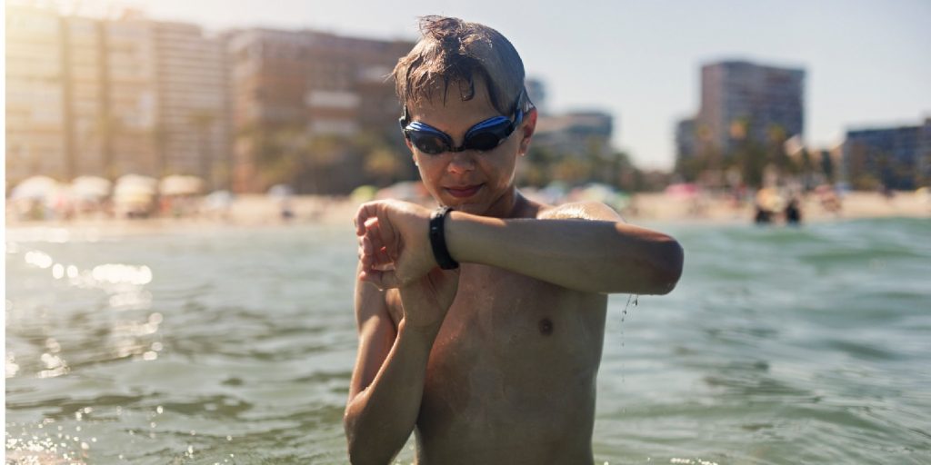 Teenage boy is swimming in the sea. He is adjusting the settings of the fitness tracker.