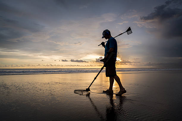 During sunset and at low tide, a Caucasian adult walks along the beach and by using a metal detector he is trying to find some hidden treasures. He is using a sophisticated device from the military and hopes to find possible gold and jewelry covered under the sand.